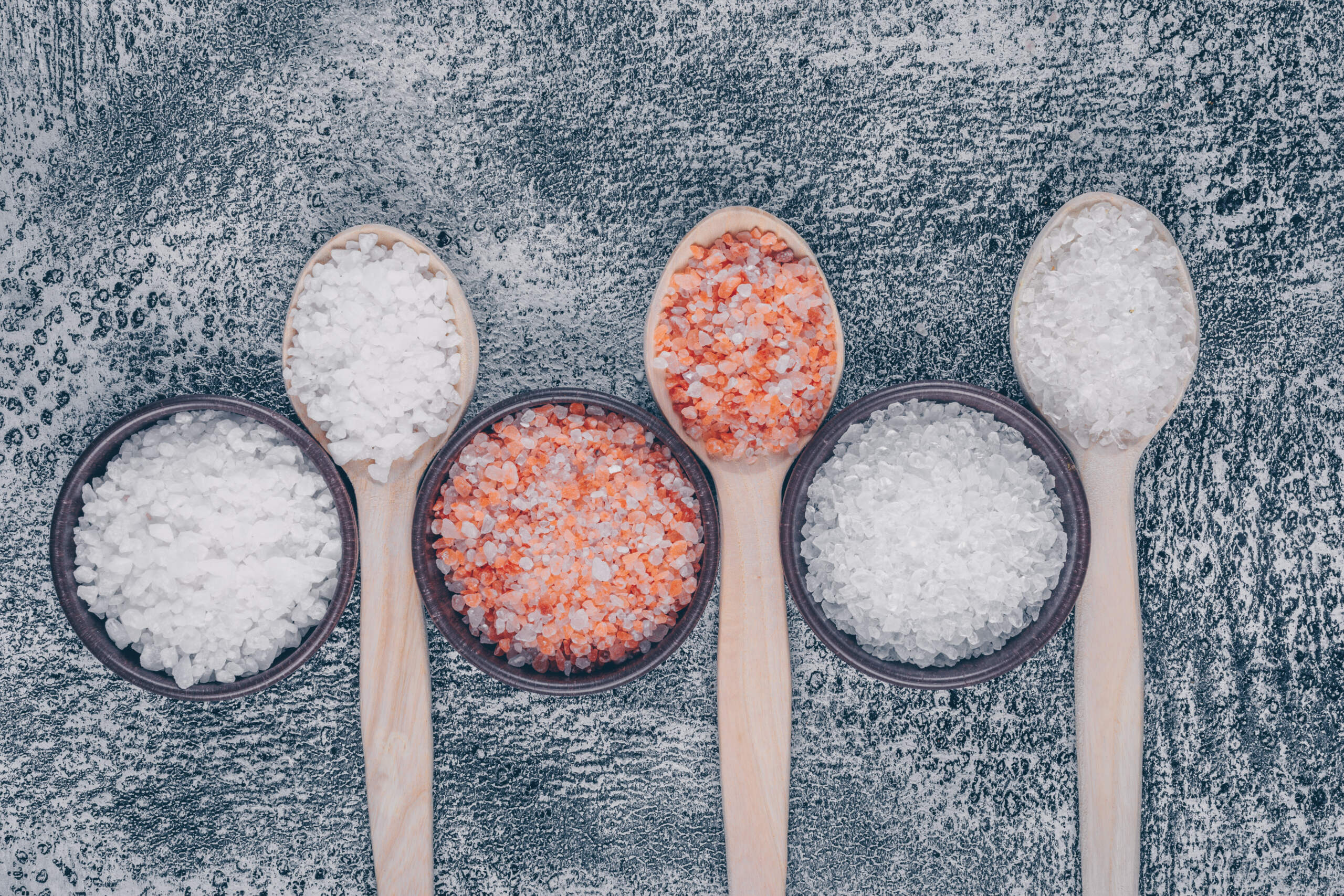 Top view sea salt in bowls and wooden spoons with himalayan salt on gray textured background. vertical
